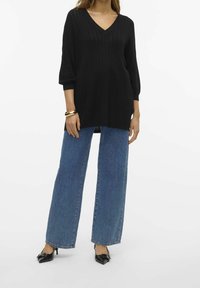 Nainen, jolla on päällään musta V-pääntie neule, leveät siniset farkut, mustat kärjetuetut korkokengät, joissa on kaarrokkeet, ja kultainen rannekoru vasemmassa ranteessa.