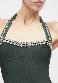 Κοντινό πλάνο ενός ατόμου που φοράει σκούρο πράσινο τοπ μπάστα, διακοσμημένο με σειρά από μεγάλα, καθαρά στρας κατά μήκος του ντεκολτέ και των τιράντων.
