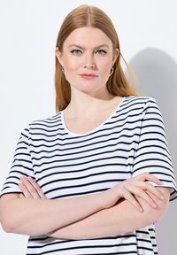 Pruhované tričko v b�ílé a černé barvě. Krátké rukávy, kulatý výstřih. Měkká tkanina s hladkou strukturou. Modelka stojí se zkříženýma rukama.