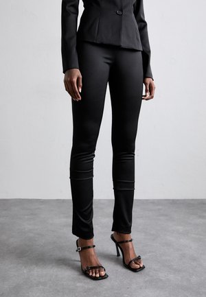 Melnas šauras bikses ar gludu tekstūru, apvienotas ar melniem siksnu augstpapēžu apaviem. Komplektā iekļauts pieguļošs žakets ar vienu pogu.