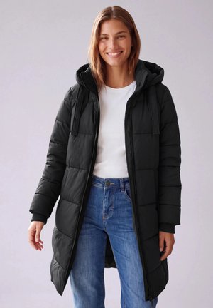 Une femme souriante porte un long manteau matelassé noir à capuche par-dessus une chemise blanche et un jean bleu, sur un fond uni.