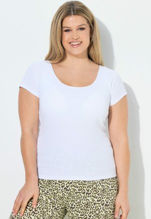 Smilende kvinde med langt blond hår iført en tætsiddende hvid kortærmet top og leopardprintede bukser mod en hvid baggrund.