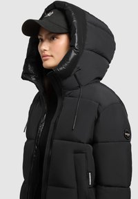 Juodas pūkinis švarkas su dideliu gobtuvu, užtrauktuku ir raišteliais. Turi priekinę kišenę ir prekės ženklą ženklelį ant rankovės.