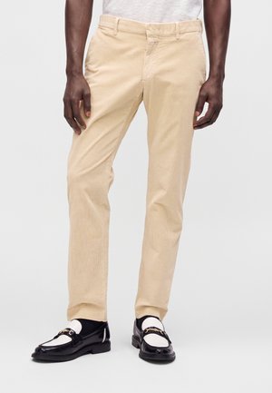 Homme portant un pantalon en velours beige et des mocassins noirs et blancs, debout devant un fond uni.