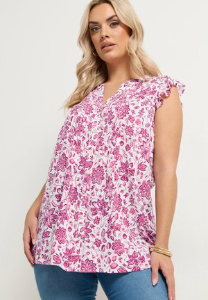 Kvinde iført hvid ærmeløs bluse med lyserødt blomsterprint, blå jeans, guldøreringe i ringform og guldbøjler mod ensfarvet baggrund.