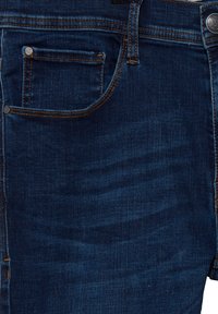 Denimjeans i en mörkblå tvätt med orange sömmar. Har en framficka, metallknapp och lapp på midjebandet. Slät textur.
