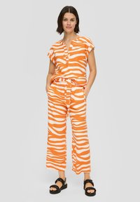 Oranje en wit gestreepte linnen twee-delige outfit met korte mouwen, een notch-kraag, wijde broekspijpen en een trekkoord in de taille. Zwarte sandalen.