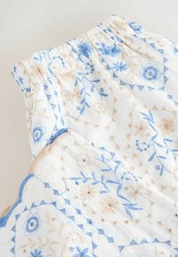 Tissu blanc avec broderie florale bleue et beige, taille élastique et bord festonné.