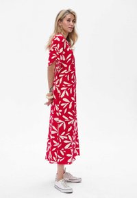 Rotes Maxikleid mit einem weißen Blättermuster, kurzen Ärmeln und einem lockeren Schnitt, kombiniert mit weißen Sneakers. Glattes Material und knöchellange Gestaltung.