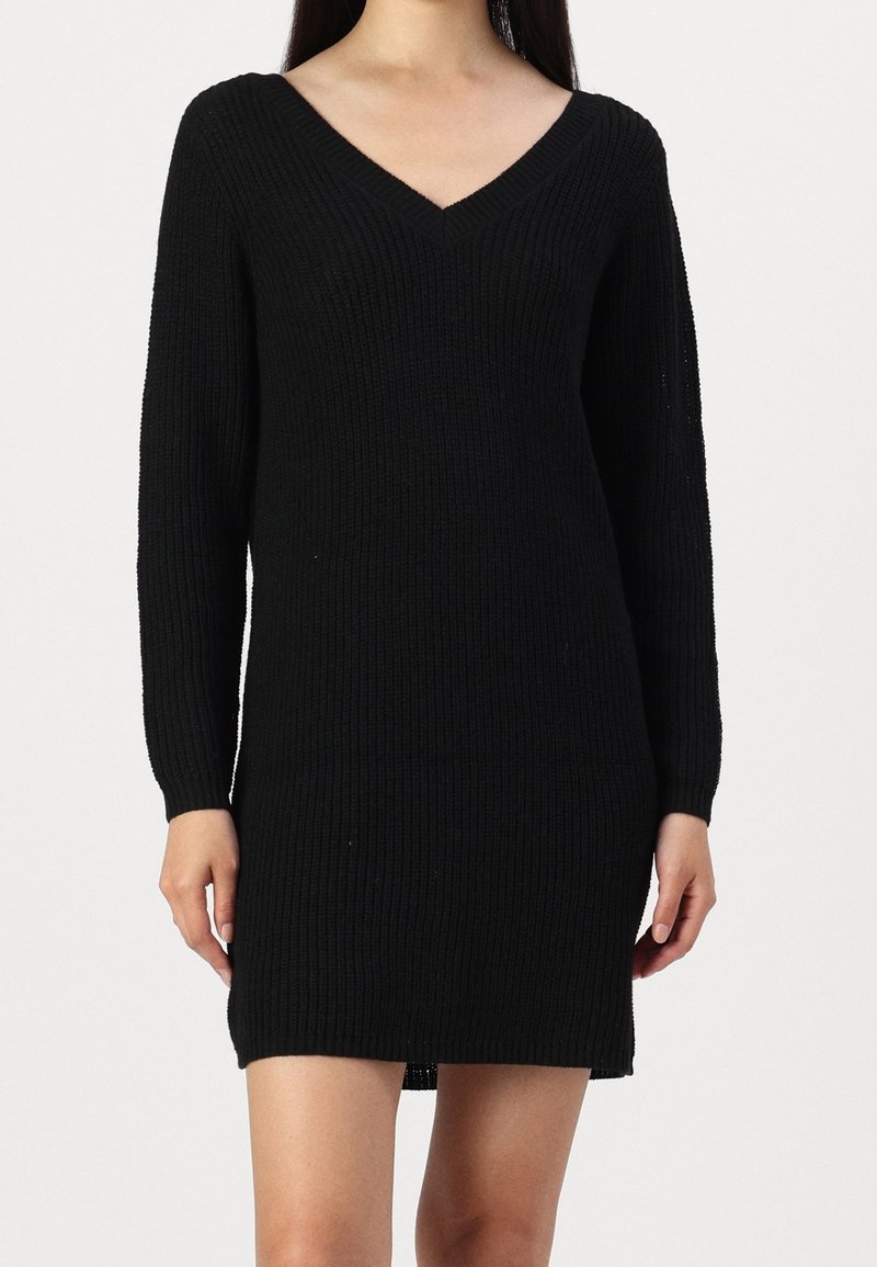 Femme portant une robe en maille côtelée noire à manches longues avec un col en V, visible des épaules jusqu'au milieu des cuisses sur un fond uni.