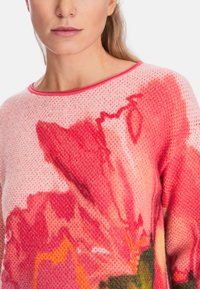 Knitted pink sweater featuring a large floral print in shades of red and orange, with a round neckline and textured fabric pattern.