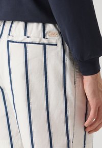 Balti dryžuoti pantaloniai su tamsiai mėlynais vertikaliais dryžiais, su užpakaline kišene ir subtiliu prekinio ženklo žymekliu, matomu ant liemenėlės.