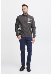 Felpa zip grigia scura con accenti rossi, caratterizzata da scritte grafiche bianche sul davanti. Abbinata a pantaloni blu navy e scarpe nere.
