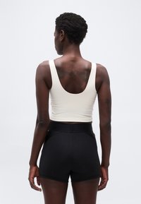 Γυναίκα με κοντά σγουρά μαλλι ά που φοράει άσπρο αμάνικο τοπ και μαύρο σορτς με ψηλή μέση, φαίνεται από πίσω μπροστά σε ένα απλό φόντο.