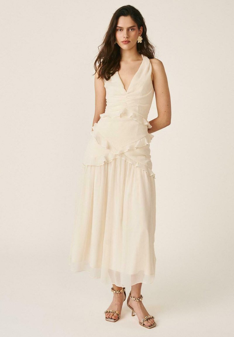 Mujer con cabello largo y oscuro, vestida con un vestido sin mangas de color crema con volantes y pendientes florales, de pie con las manos detrás de la espalda.