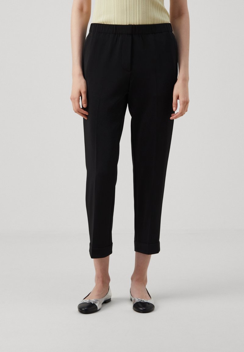 Pantalon noir fuselé avec une ceinture élastique, présentant une texture lisse et des ourlets retroussés, associé à des ballerines métalliques et noires.