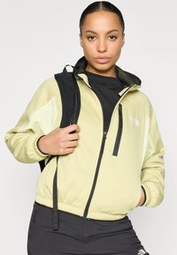 Jachetă verde deschis cu fermoar și accente negre, cu un model texturat pe umeri și manșete elastice, purtată cu un top negru.