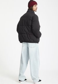 Zwarte gewatteerde jas met een hoge kraag en een gestikt ontwerp; gecombineerd met lichtblauwe wijde jeans en zwarte sneakers. Rode en zwarte gestreepte beanie.