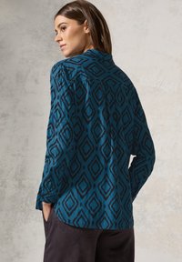 Camicia teal con motivo a rombi neri, maniche lunghe e colletto, indossata con pantaloni scuri. Texture liscia, design casual.
