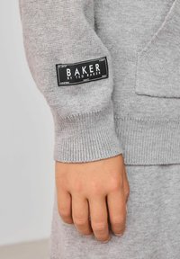 Sivý pletený sveter s rebrovanými manžetami, s čiernou nášivkou "BAKER BY TED BAKER." Tkanina je jemná a textúrovaná.