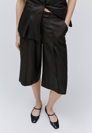 Sorte, teksturerede, vidde-bukser med korte ben, kombineret med sorte Mary Jane-flats, båret med den ene hånd i lommen mod en ensfarvet baggrund.