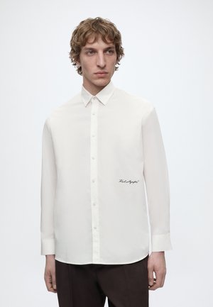 Jeune homme aux cheveux bouclés châtain clair portant une chemise blanche boutonnée avec une écriture noire près de la taille et un pantalon brun foncé.