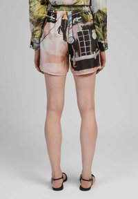 Femme portant un short imprimé et une chemise à manches longues, debout pieds nus sur un sol gris clair, tournant le dos à l'appareil photo.