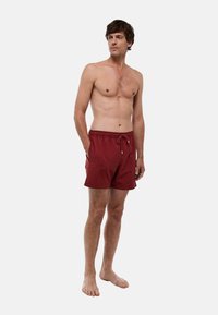 Shorts de bain rouges avec un design texturé, taille élastique et cordon de serrage. Dotés de poches latérales. Portés pieds nus sur un fond uni.