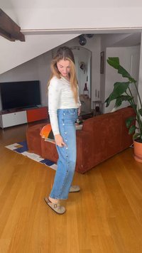 Femme aux cheveux longs portant un haut blanc et un jean bleu, pointant du doigt les boutons le long de la couture latérale, debout dans un salon.