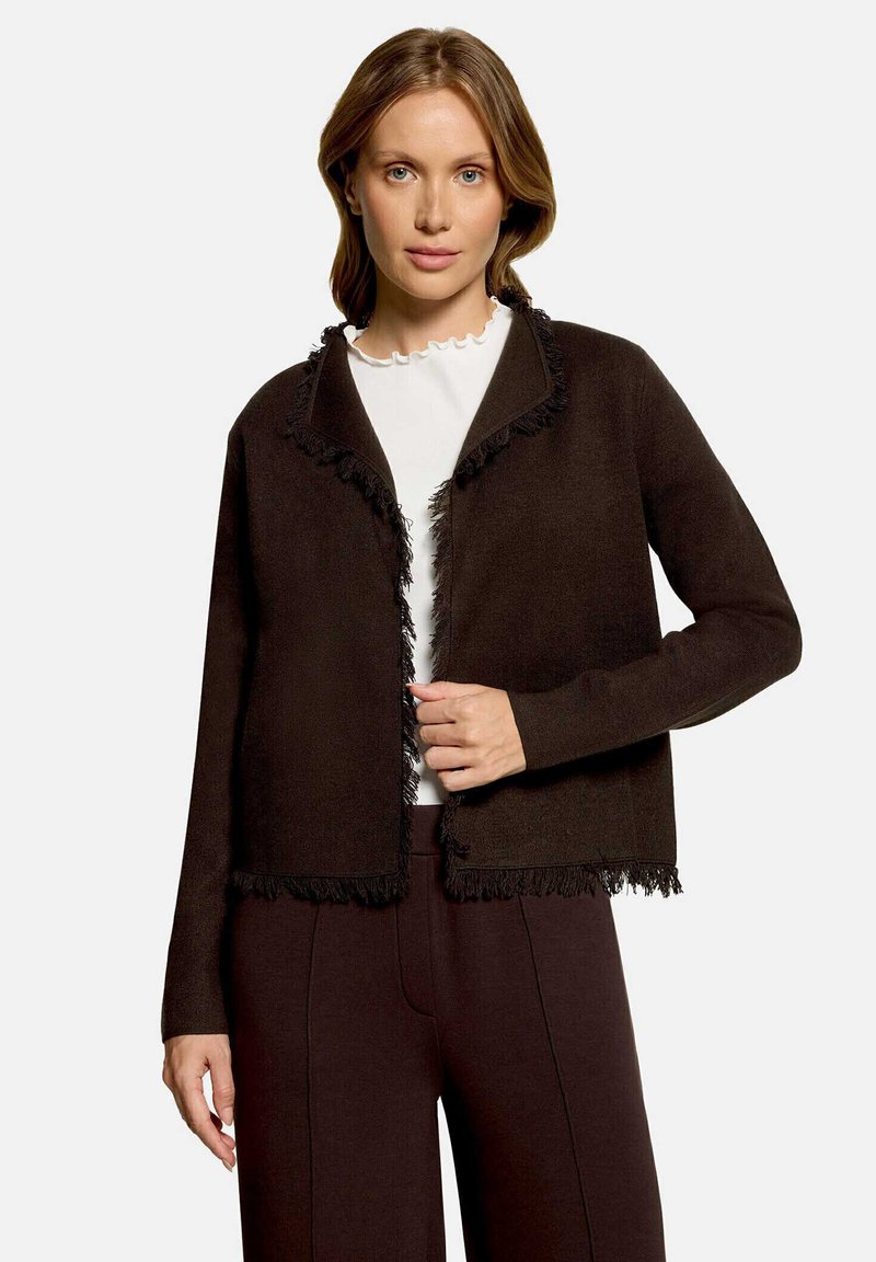 Femme portant une veste marron foncé à franges sur un haut blanc avec un col à volants et un pantalon marron foncé assorti, debout devant un fond uni.