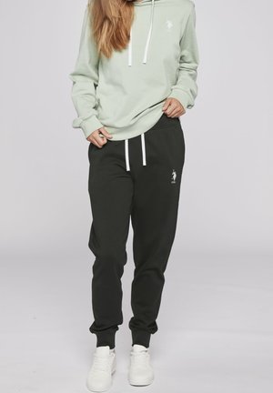 Frau trägt hellgrünen Kapuzenpullover und schwarze Jogginghosen mit weißen Kordeln sowie weiße Turnschuhe, steht vor einem einfarbigen Hintergrund.