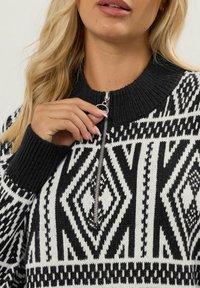 Schwarz-weißer, gemusterter Strickpullover mit hohem Kragen und halbem Reißverschluss. Hat geometrische Designs und strukturierte Rippbündchen.