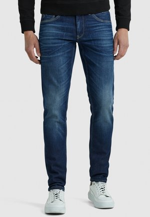Mann in dunkelblauen Slim-Fit-Jeans mit ausgebleichten Details und weißen Sneakers, stehend vor einem schlichten hellen Hintergrund.
