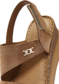 Brown suede wedge sandal with adjustable strap, featuring a smooth tan insole and a metallic logo accent on the side.