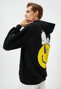 Sudadera negra con un gran gráfico de una cara sonriente amarilla en la parte posterior. Con capucha ajustable y puños acanalados. Textura de tela suave.