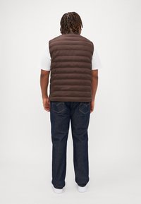 Brun puffer väst med horisontella sömmar, över en vit skjorta. Buren med mörkblå jeans och vita sneakers, sedd bakifrån.