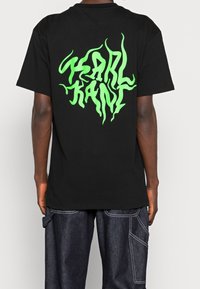 Černé bavlněné tričko s velkým, živým zeleným grafickým designem na zádech, na kterém je nápis "KANI" v tekoucím, vlnitém písmu.