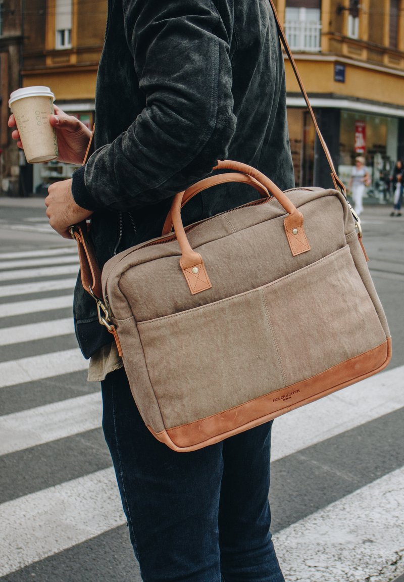Canvas and leather messenger bag in beige with brown leather accents, zip closure, and front pocket. Adjustable shoulder strap shown.
