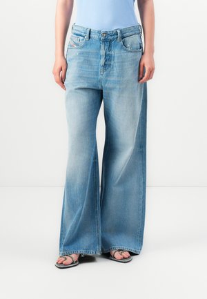 Person wearing light blue wide-leg jeans and strappy black sandals with thin heels, standing against a plain white background.