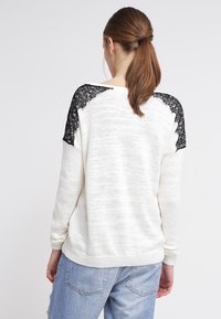 Femme avec une queue de cheval brune portant un pull blanc en tricot avec des épaules en dentelle noire et un jean bleu clair, vue de dos.