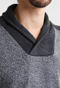 Grauer Pullover mit strukturiertem Muster, Schalkragen und kontrastierendem dunklem Kragenmaterial, das eine Mischung aus Weichheit und Wärme hervorhebt.