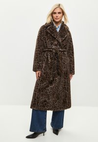 Leopardikuvioitu tekoturkistakki, jossa on vyötäröllä vyö, kaksirivinen malli, suuret kaulukset ja pitkät hihat; päällä leveälahkeisten farkkujen kanssa.