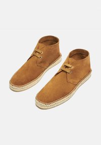Botas de tobillo de ante marrón con suelas de yute tejido en beige y cierre de ojetes. El diseño presenta una punta redondeada y acentos de costura mínimos.