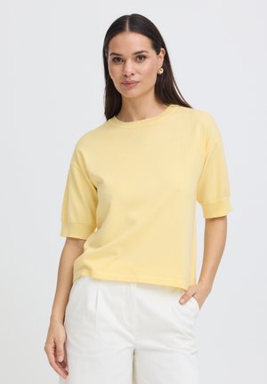 Kvinde med langt mørkt hår iført gul kortærmet sweater og hvide bukser, stående med den ene hånd i lommen mod en ensfarvet baggrund.