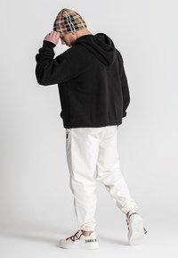 Sudadera negra con capucha, pantalones de pana blancos, gorro a cuadros y zapatillas blancas con acentos rojos. Diseño simple y casual con un corte holgado.