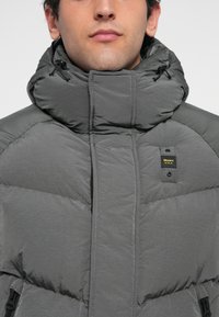 Chaqueta acolchada gris con cuello alto, textura acolchada, cremallera frontal y etiqueta de marca en el pecho. Diseñada para ofrecer calor y comodidad.