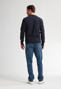 Marineblauwe trui met een ronde halslijn, lange mouwen en een geribde zoom, gecombineerd met lichtblauwe jeans en witte sneakers, gezien van achteren.