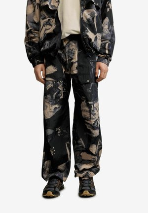Személy fekete és bézs absztrakt mintás cargo nadrágot és hozzá illő dzsekit visel, fekete és barna túracipővel, kezei lazán állnak.