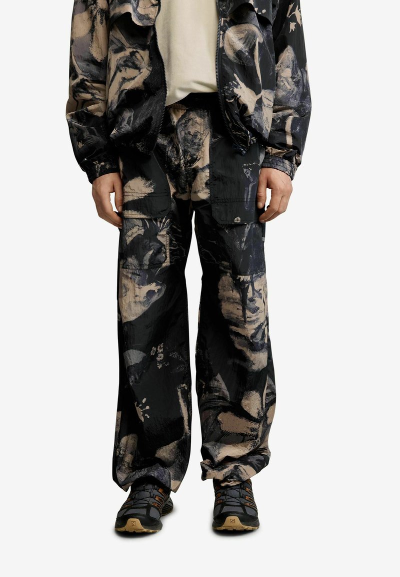 Asmuo dėvi juodos ir smėlio spalvos abstraktaus rašto krovinių kelnes bei derančią striukę, su juodais ir rudos spalvos žygių batais, stovėdamas atsipalaidavusiomis rankomis.