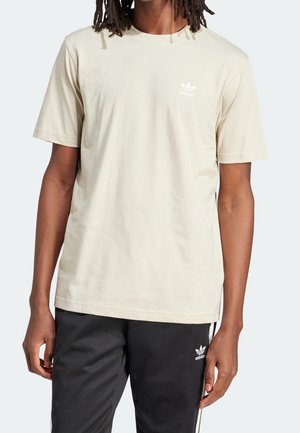 Mężczyzna w beżowej koszulce Adidas i czarnych spodniach dresowych Adidas z białymi paskami po bokach, stoi z rozluźnionymi ramionami.